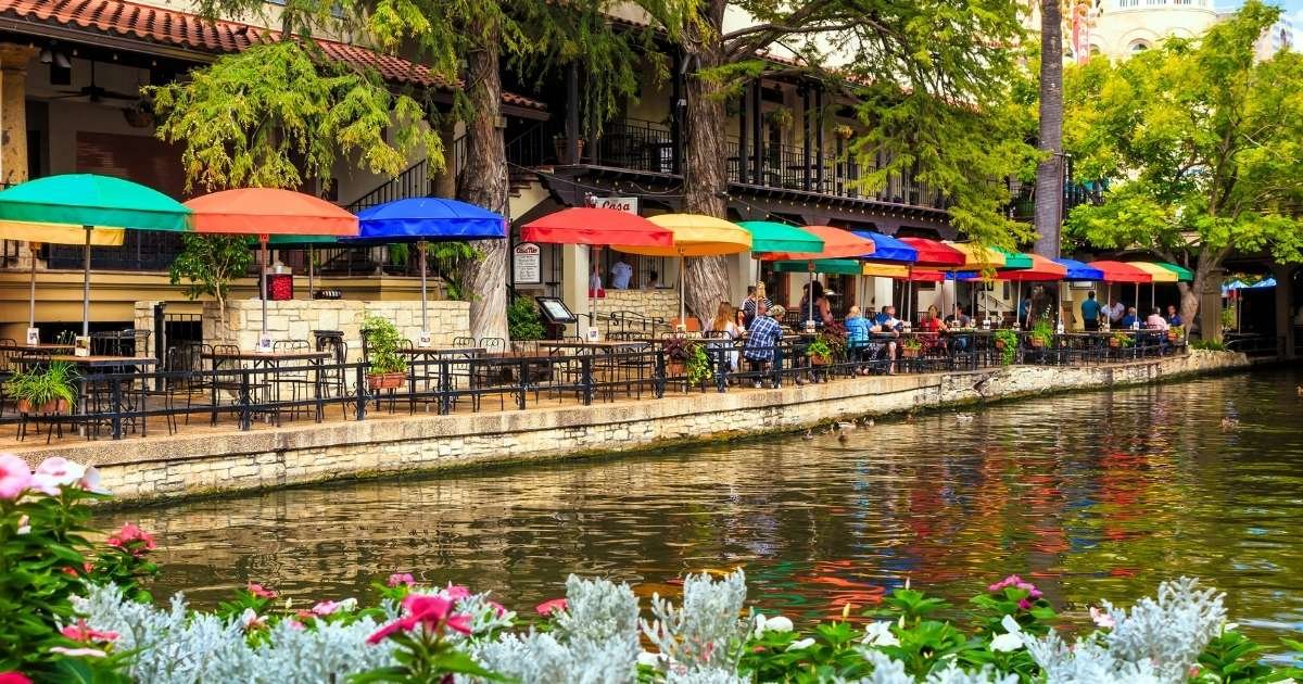 Waterfront shopping and dining area. Umbrella shades in many colors cover tables where people are eating and chatting. San Antonio is a great place for a date.