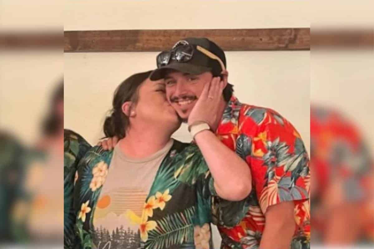 Logan and Jourdan posing for a photo in vacation wear. She is planting a kiss on his cheek while he smiles toward the camera.