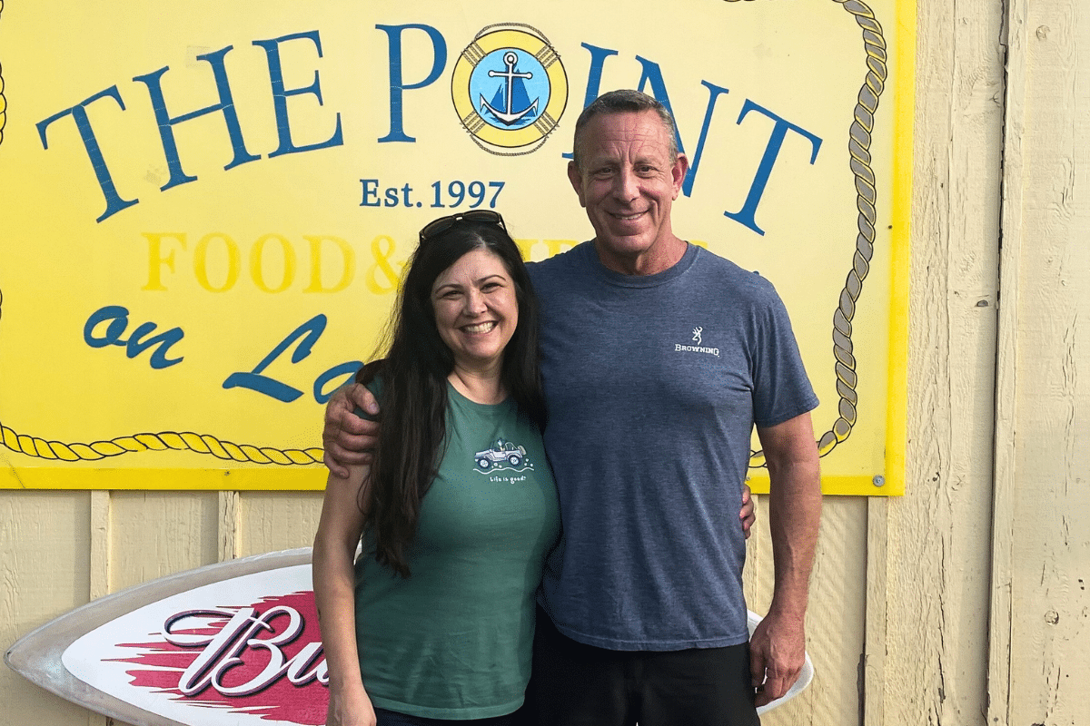 Matchmaking success story couple standing in front of a surf board and yellow sign that reads "The Point"