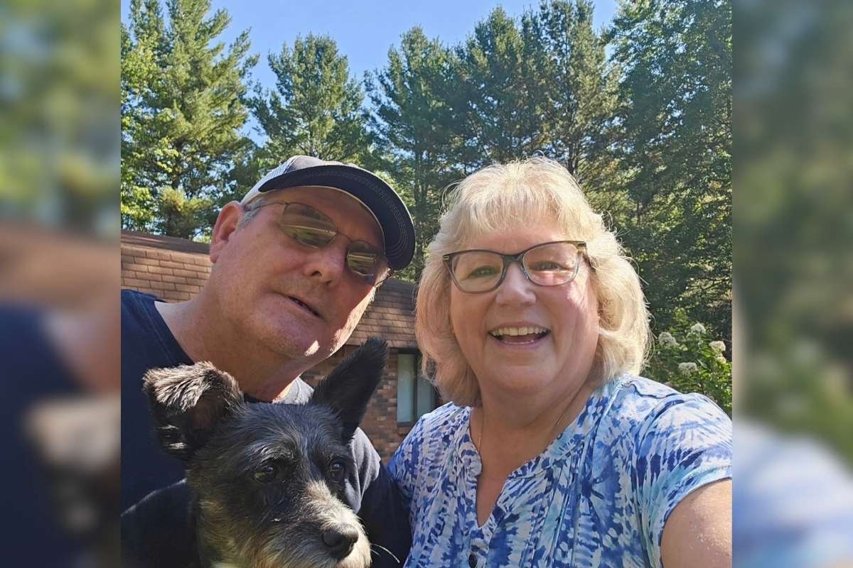 Couple in the 60s smiling with their dog in the sunshine with green trees in the background